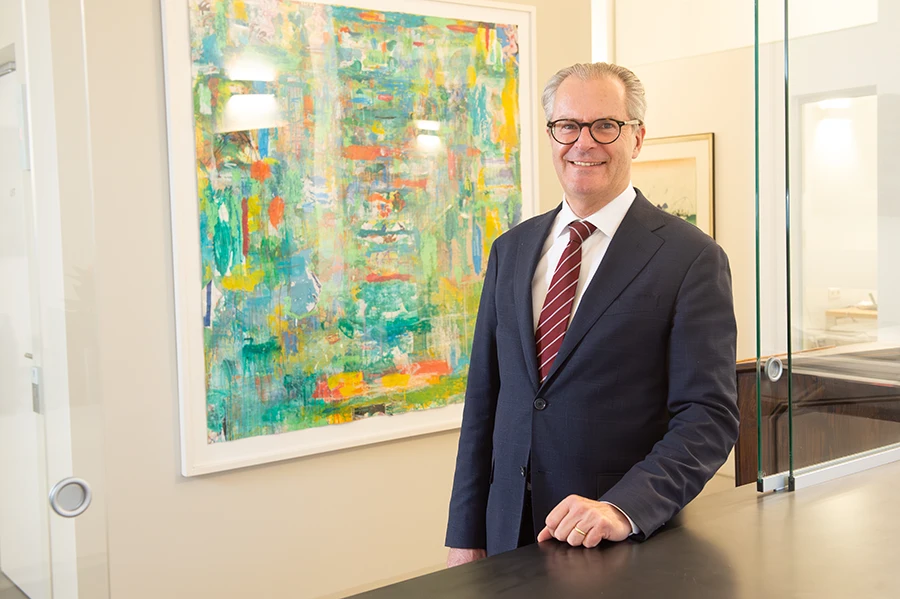 Notar Dr. Erfried Bäck, ein grauhaariger Mann mit Brille, steht im Büro an einem Tisch und blickt lächelnd in die Kamera.