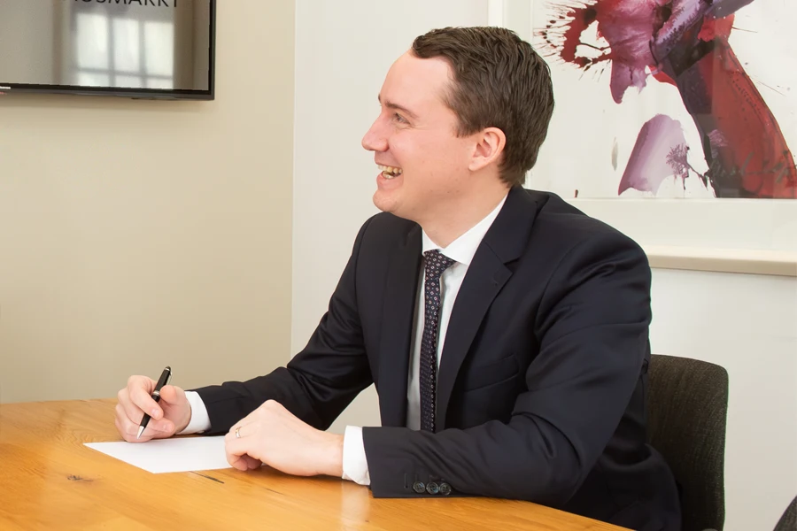 Notar Mag. Michael Niederegger, ein Mann mit kurzen dunklen Haaren, sitzt im Büro an einem Tisch und schreibt lächelnd auf ein Dokument.